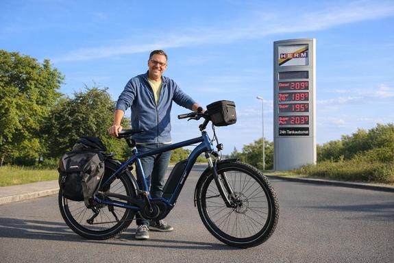 Tank-Rabatt beim E-Bike-Kauf in Lauda Mitarbeiter mit E-Bike vor Tankstellen-Preistafel in Lauda – Tank-Rabatt beim E-Bike-Kauf